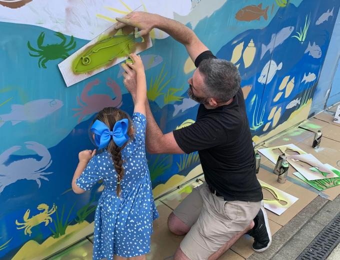 A man helping a little girl spray paint a fish on the wall.