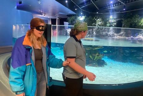 Two of The Deep's Guides walking past Lagoon of Light, one is blindfolded and the other is using the Guiding Arm technique to guide the blindfolded person around the aquarium.