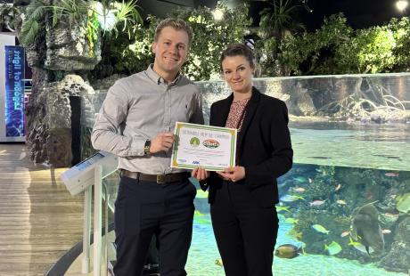 Chris and Charlotte Elliott stood smiling holding their Sustainable Palm Oil champion certificate in front of The Deep's Lagoon of Light exhibit.